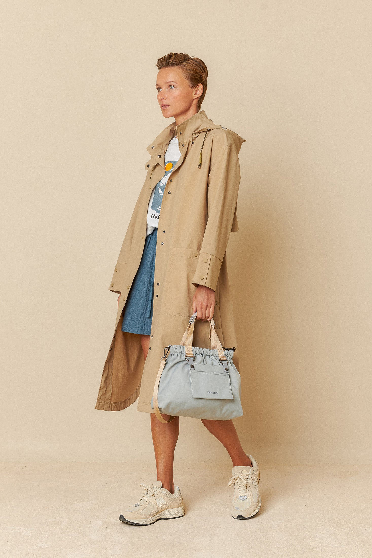 Woman wearing a beige trench coat holding a light blue handbag against a beige background